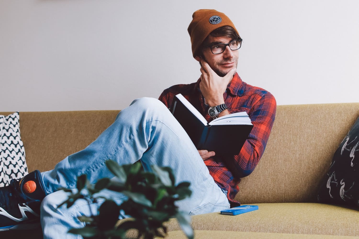 Man with a beanie and glasses with crossed legs on a couch with a book